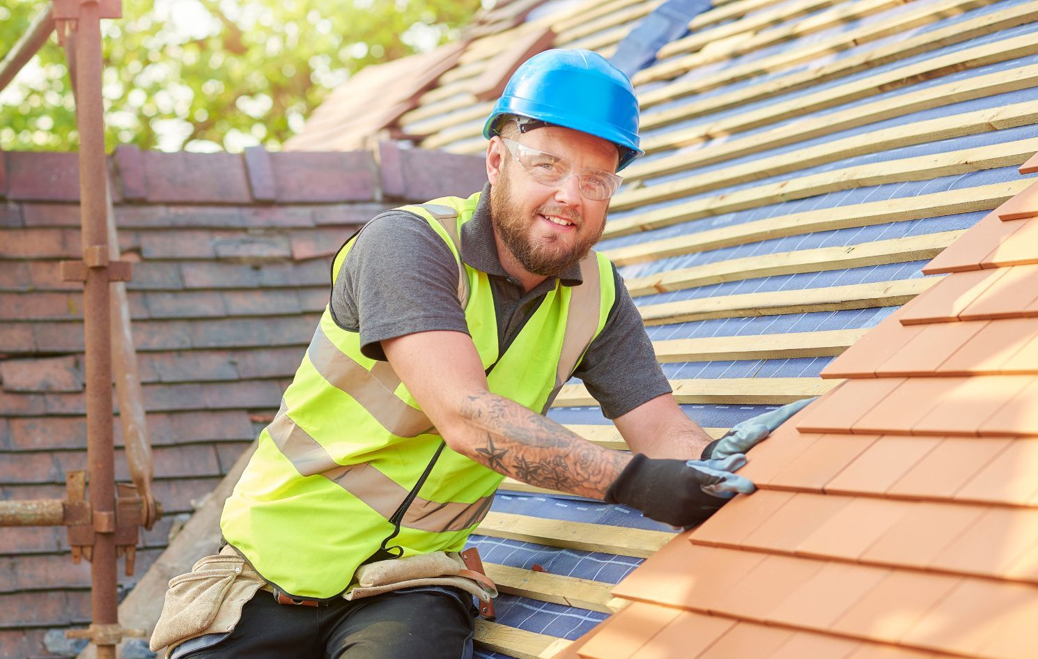 Roofer on a roof, wearing safety gear, smiling at the camera while working with tiles.