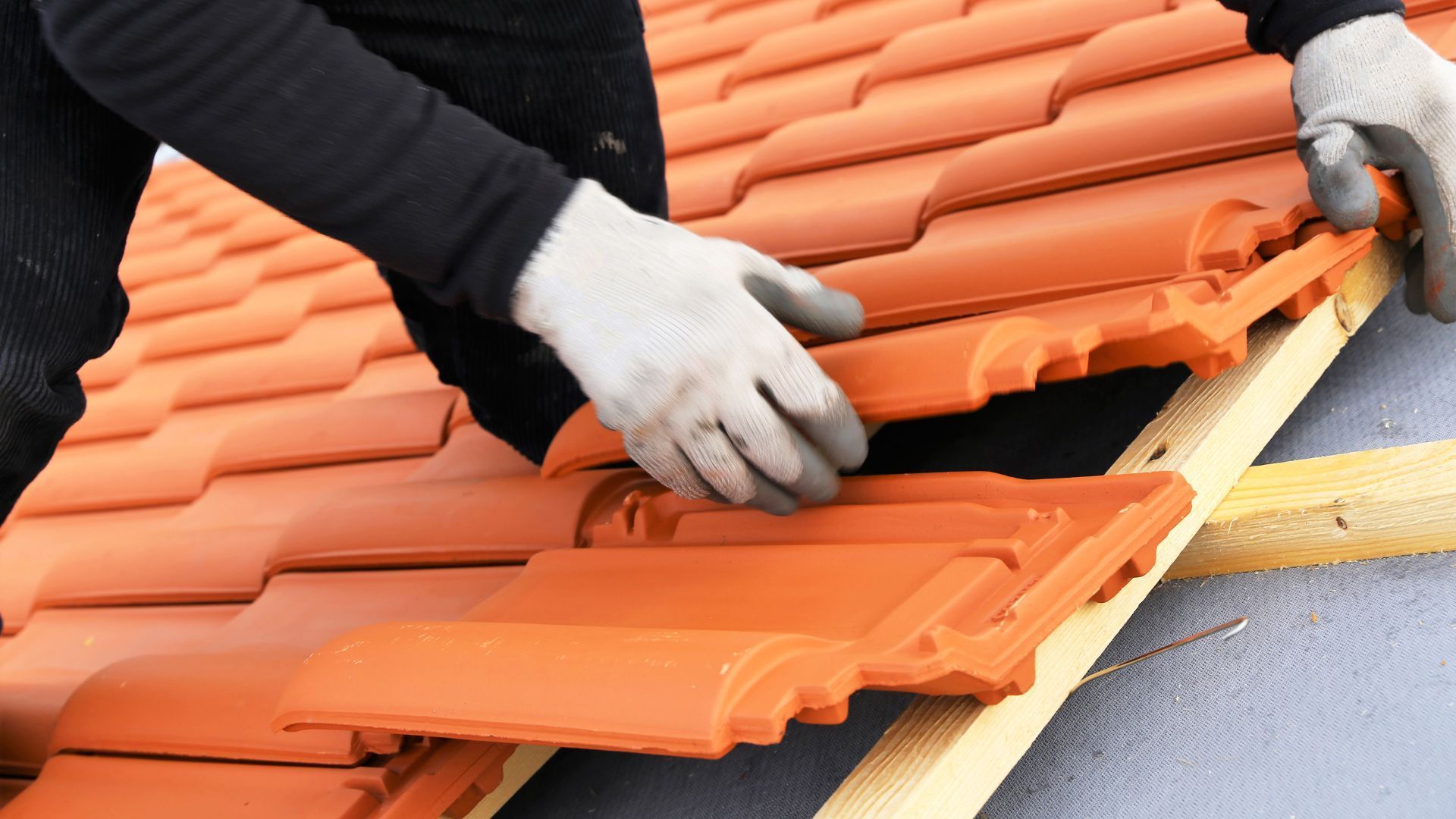 A person with gloves placing a terracotta roof tile on a wooden frame.