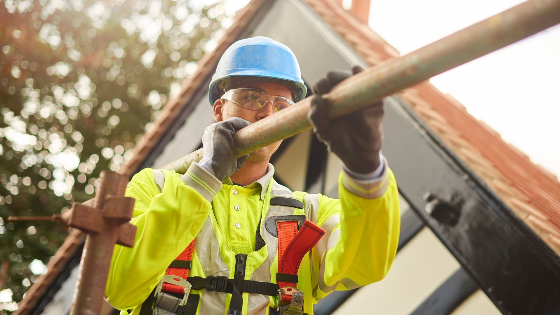 Construction worker on scaffolding.  Wearing safety gear, he holds a metal pole against a roof.