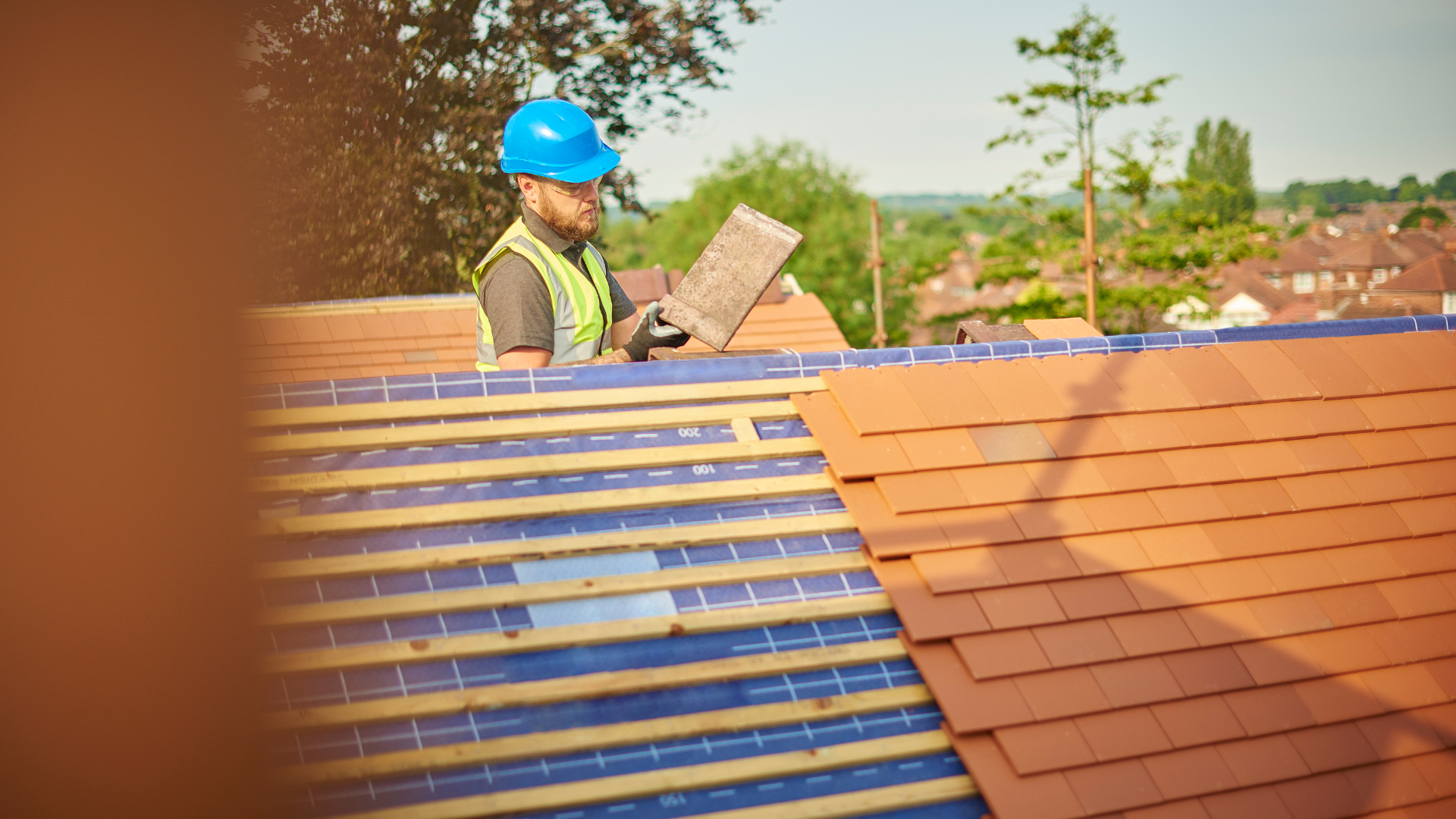 Roofer in blue hard hat inspecting roof tiles on a sunny day.