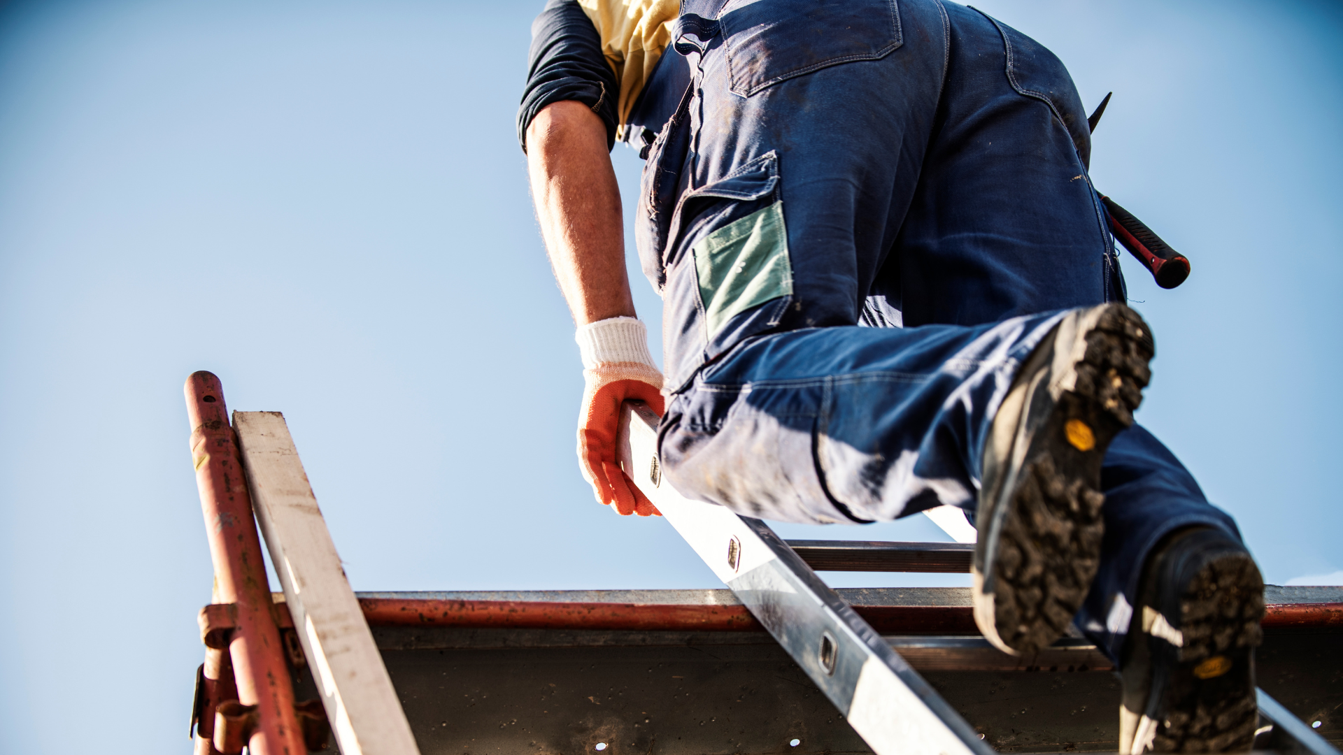 Person in work clothes climbing a ladder onto a roof, against a blue sky.