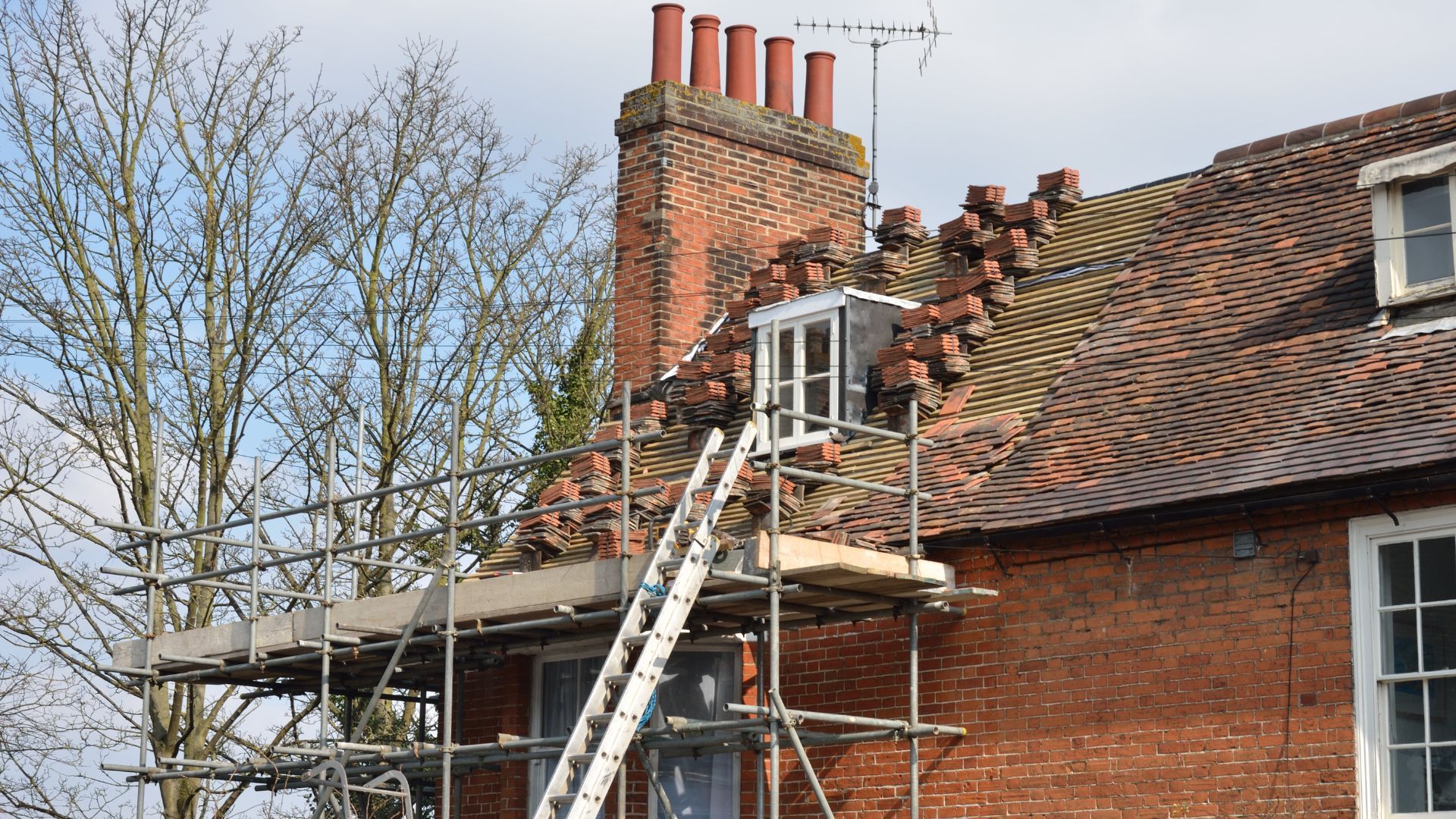 Brick house roof under repair with scaffolding and scattered tiles; chimney at top.