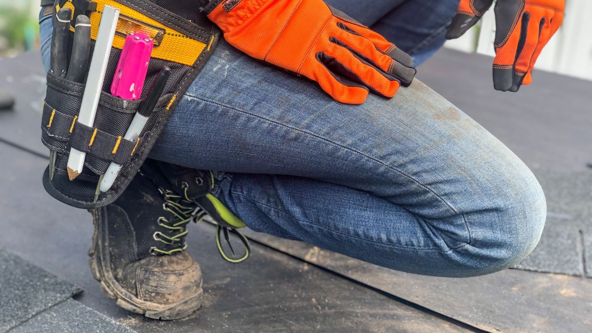 Person in work boots and gloves squats on a roof, tool belt visible.