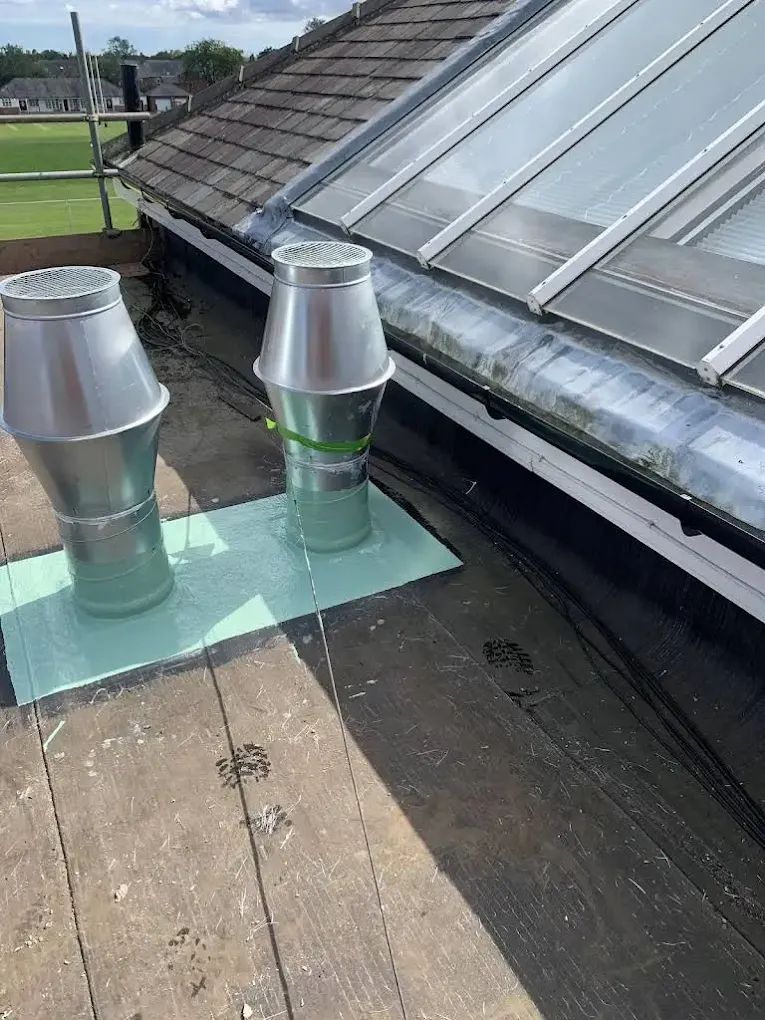 Two metal vent pipes on a roof, beside a skylight. Green insulation pads and black tar visible.