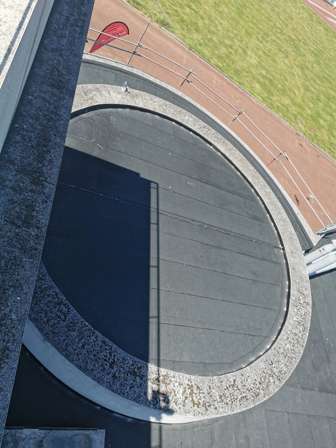 View from above of a circular asphalt area surrounded by concrete, next to a running track and grass field.