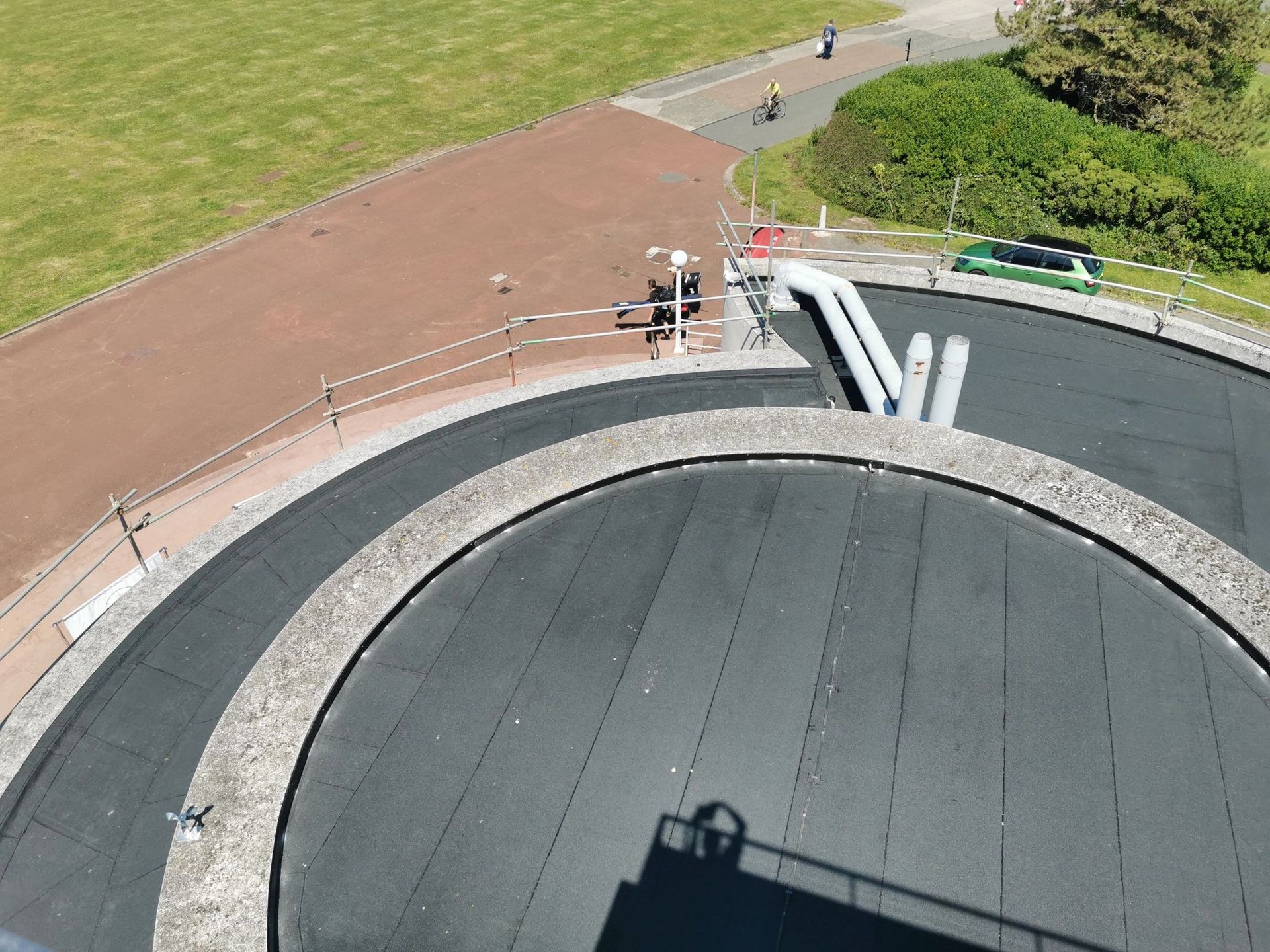 Overhead view of a circular rooftop with dark asphalt and a concrete border, next to a walkway and green grass.