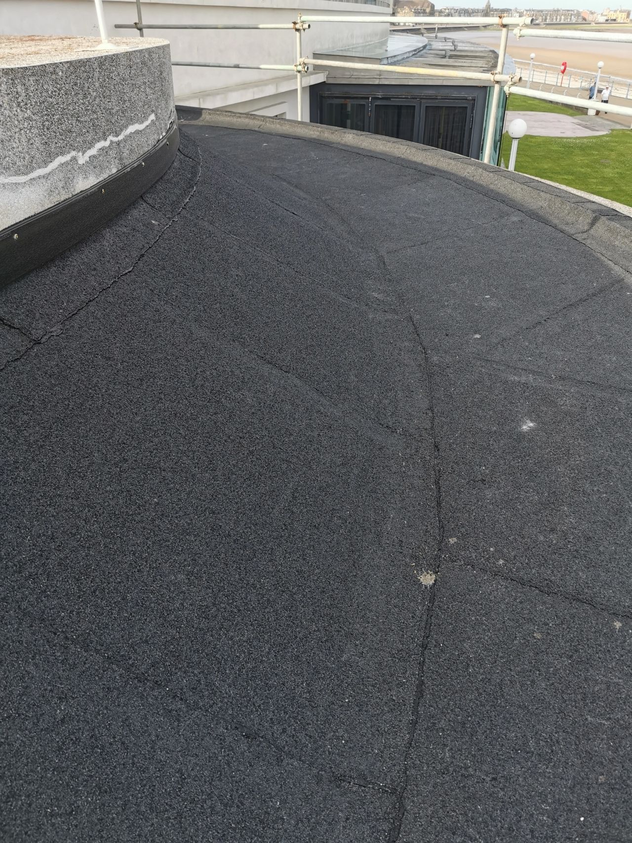 Black, textured, flat roof with slight cracks and curved edge, with a building and cityscape in the background.