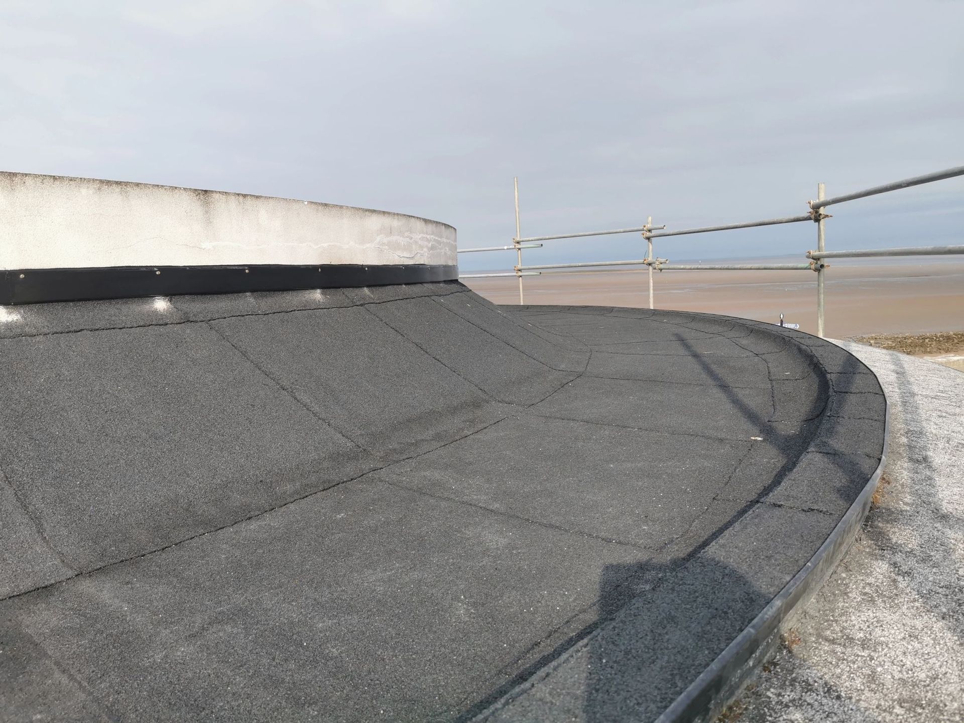 A curved, flat, asphalt roof edge with a white ledge, beach and scaffolding visible in the background.