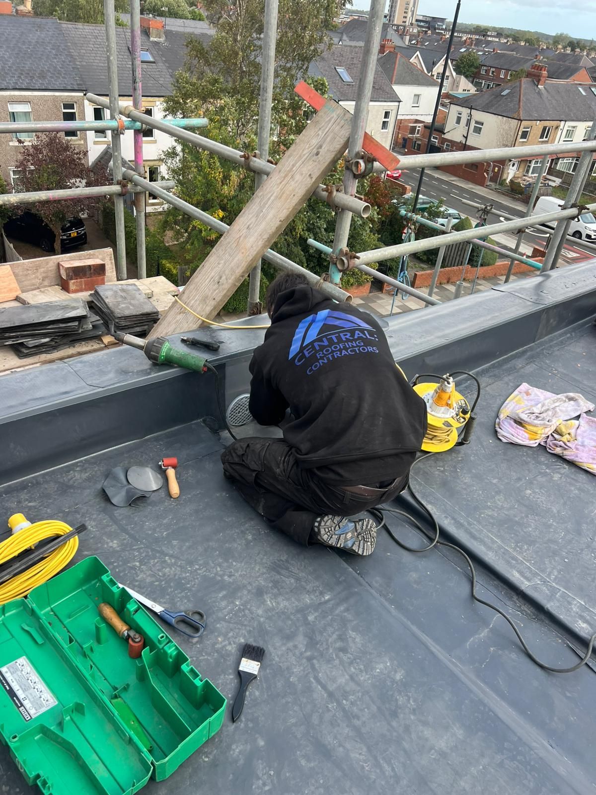 A flat roof with a red-brown surface, brick wall, and a slate tile roof visible in the background.