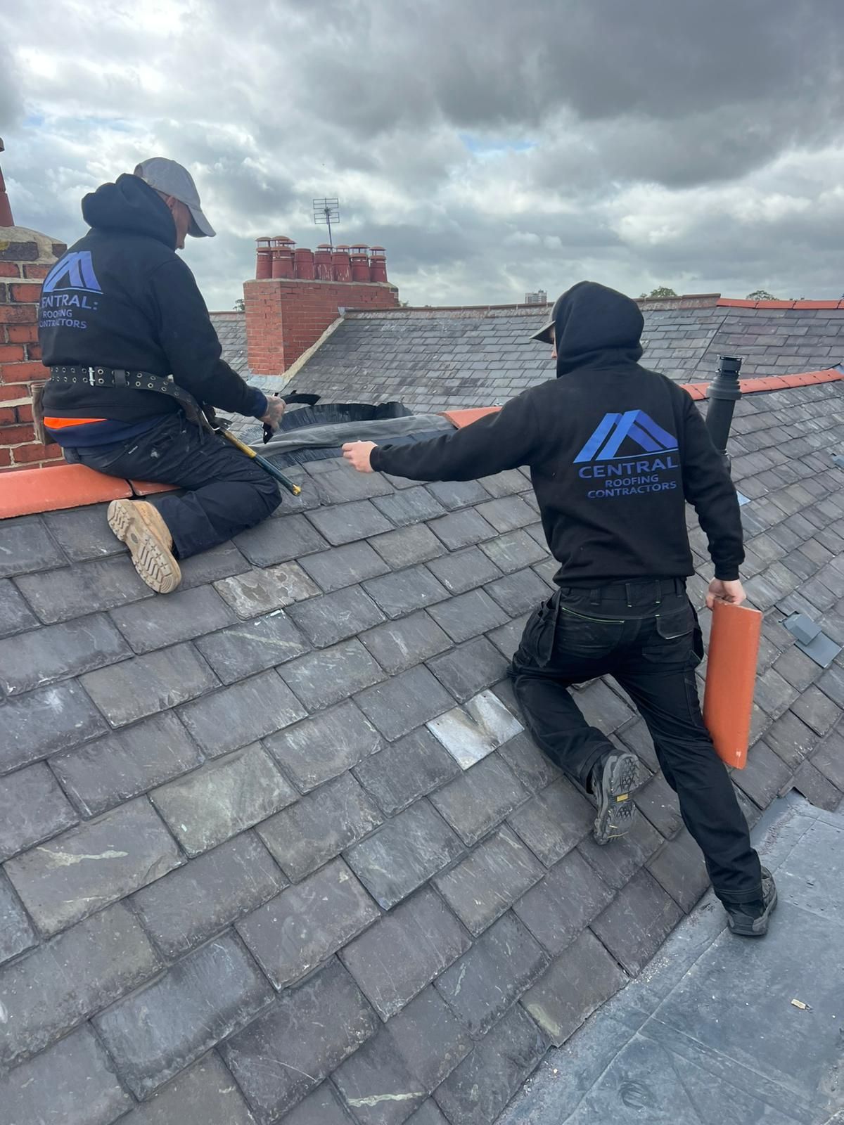 Two roofers wearing black hoodies working on a slate roof, cloudy sky overhead.
