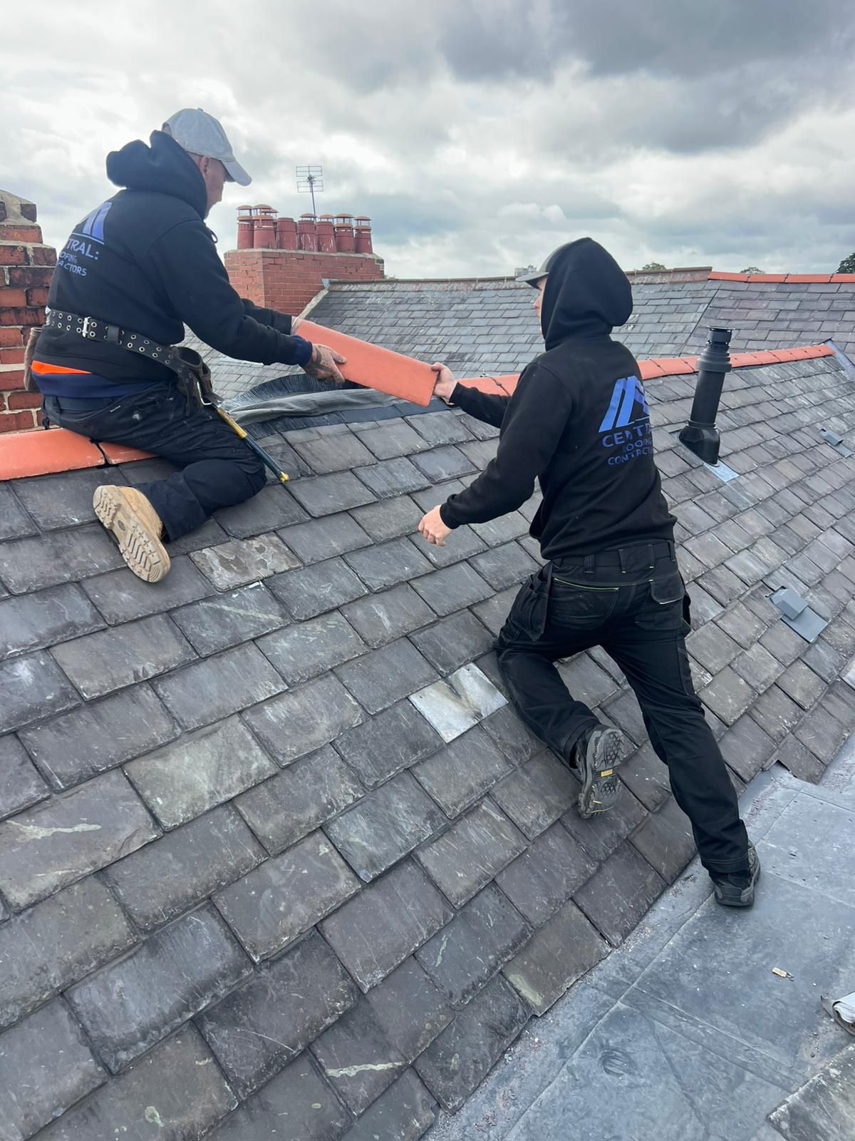 Two roofers on a gray slate roof, one handing the other a piece of red material. Overcast sky.