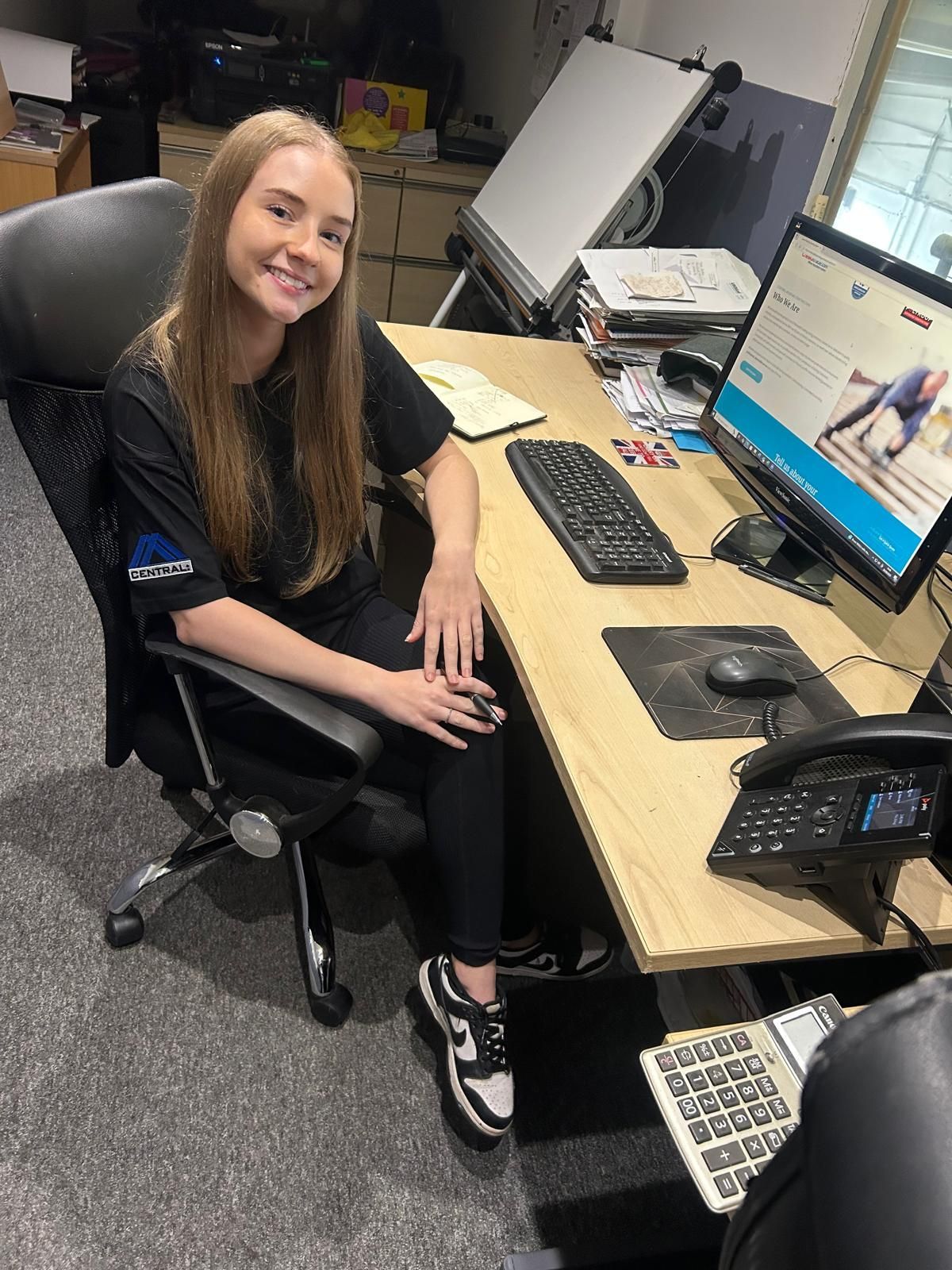Woman sitting at a desk, smiling. Wearing black clothes and sneakers. Office setting with computer and phone.