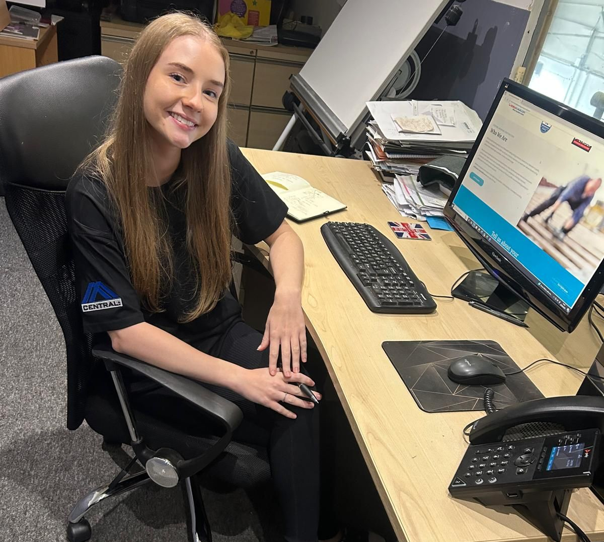 Woman with blonde hair smiles at a desk in an office. She has a computer, phone, and keyboard in front of her.