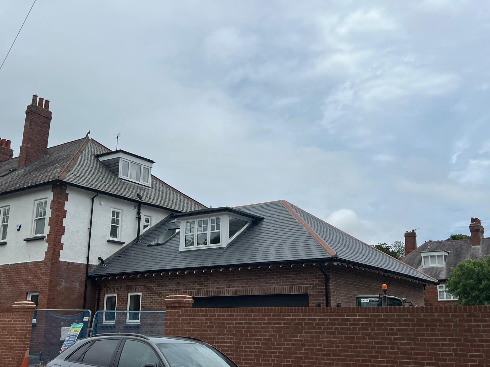 Brick buildings with slate roofs and dormer windows, behind a brick wall under a cloudy sky.