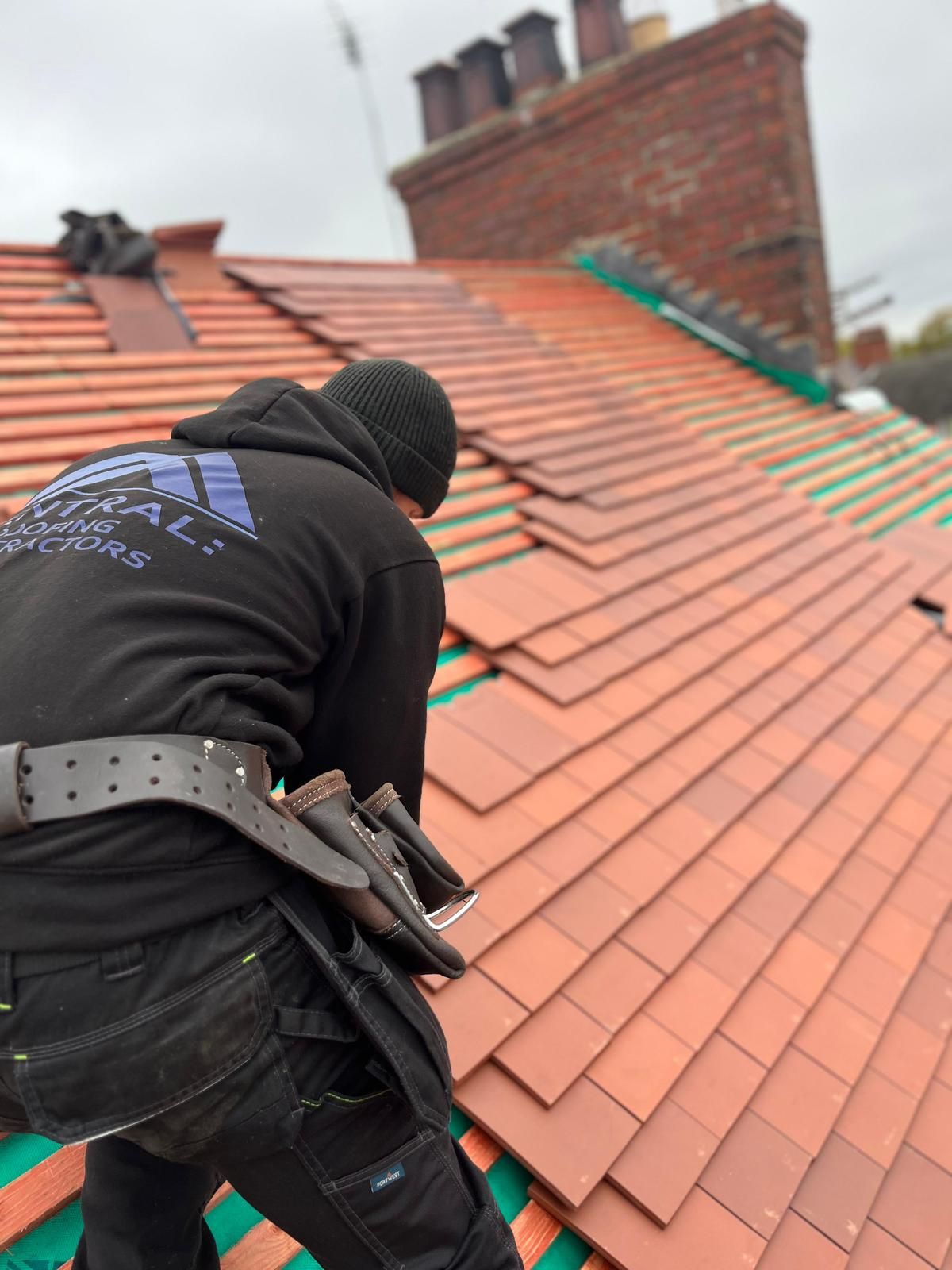 Roofer installing red tiles on a pitched roof, working near a brick chimney under an overcast sky.