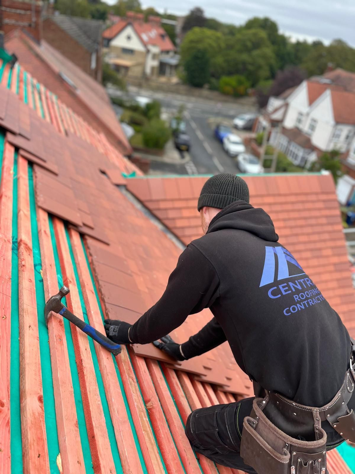 Roofer in black hoodie hammering tiles on a roof with a residential street in the background.