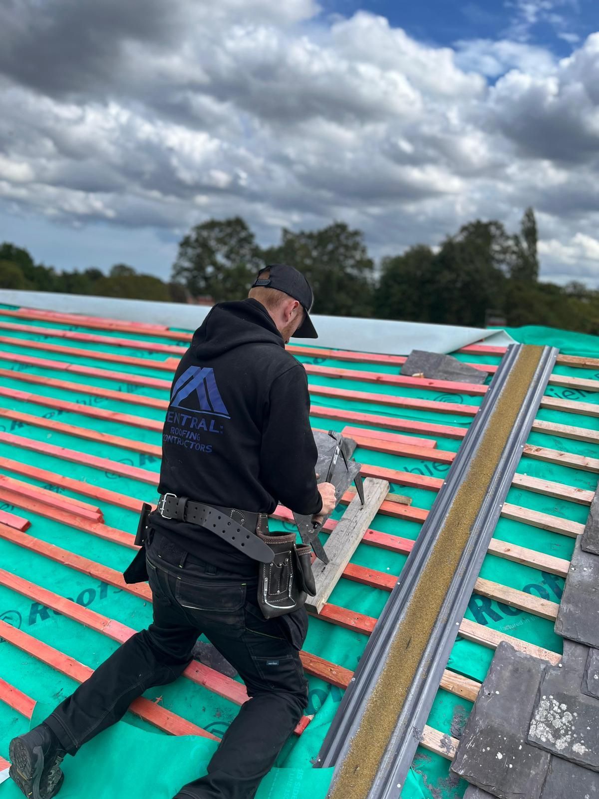 Two roofers wearing black hoodies working on a slate roof, cloudy sky overhead.