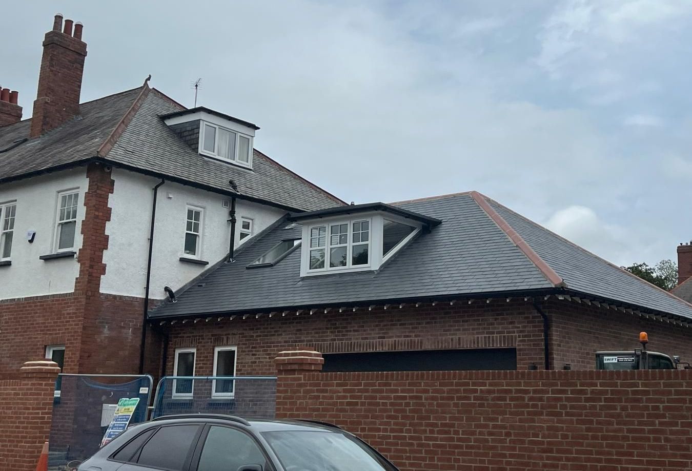 Building with a slate roof, brickwork, and dormer windows. Grey car parked in front of a brick wall.