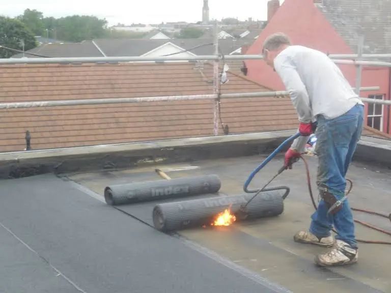 Roofer using a torch to heat and seal a roll of roofing material on a flat roof.
