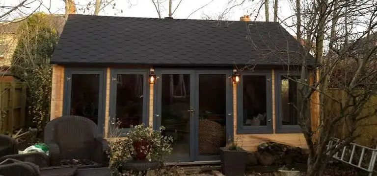 A small, dark gray-roofed garden shed with large windows and doors, surrounded by plants and trees.