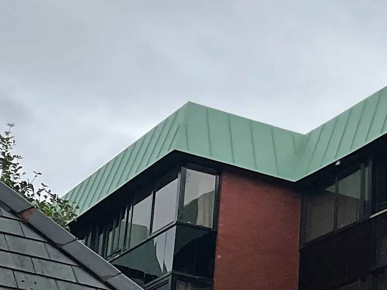 Green metal roof on a modern building with a red brick facade and large windows under a cloudy sky.