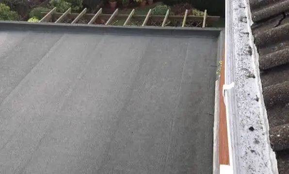 Dark flat roof next to a dirty gutter and angled roof tiles; a wooden structure is in the background.