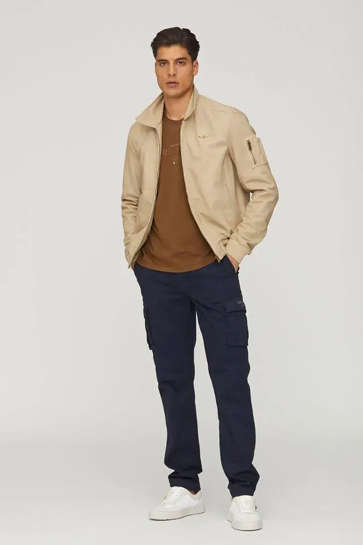 Un uomo che indossa una giacca marrone chiaro e pantaloni blu è in piedi con le mani in tasca.