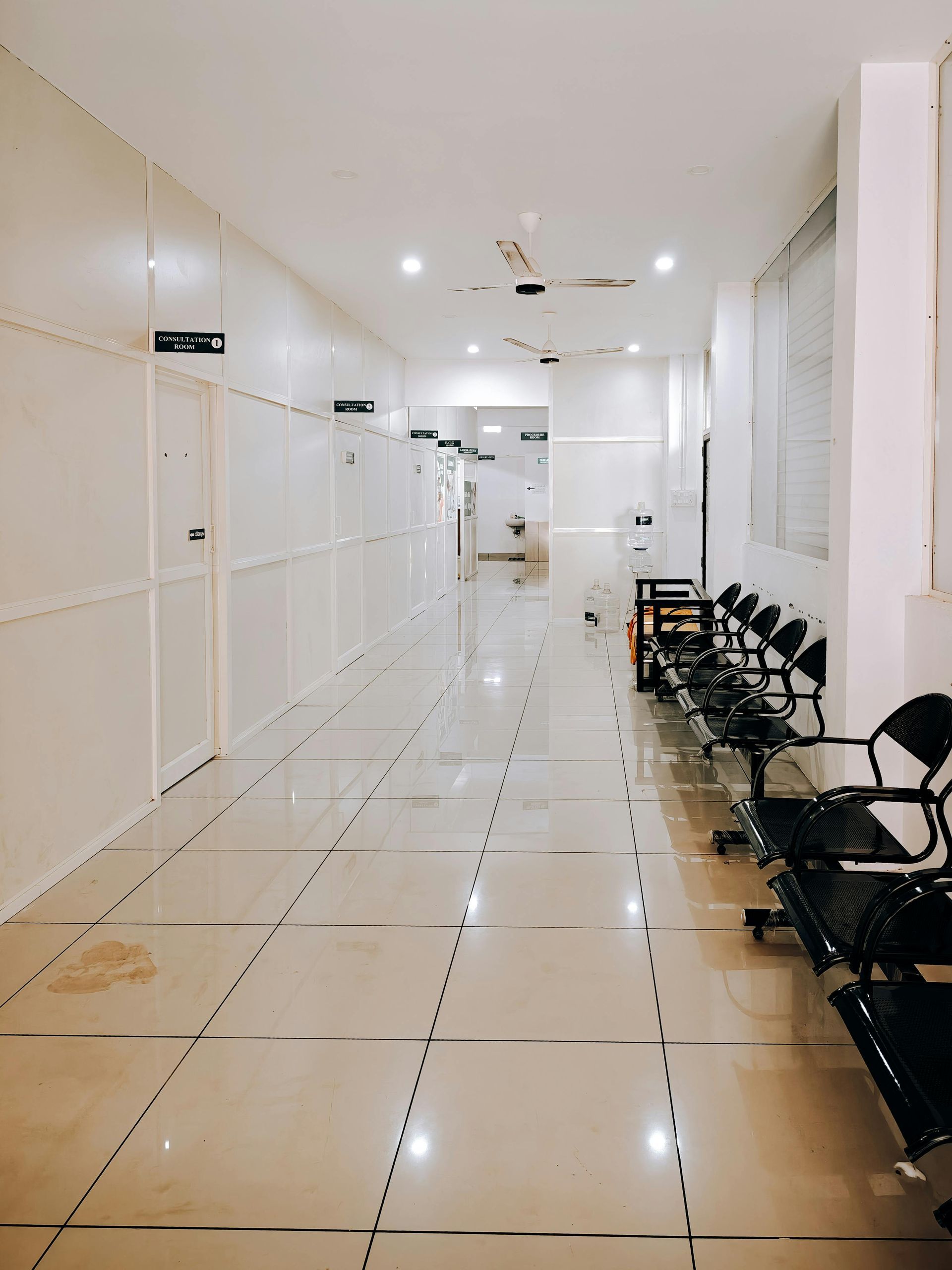 Bright white hallway with tiled floor and rows of black chairs along the right wall
