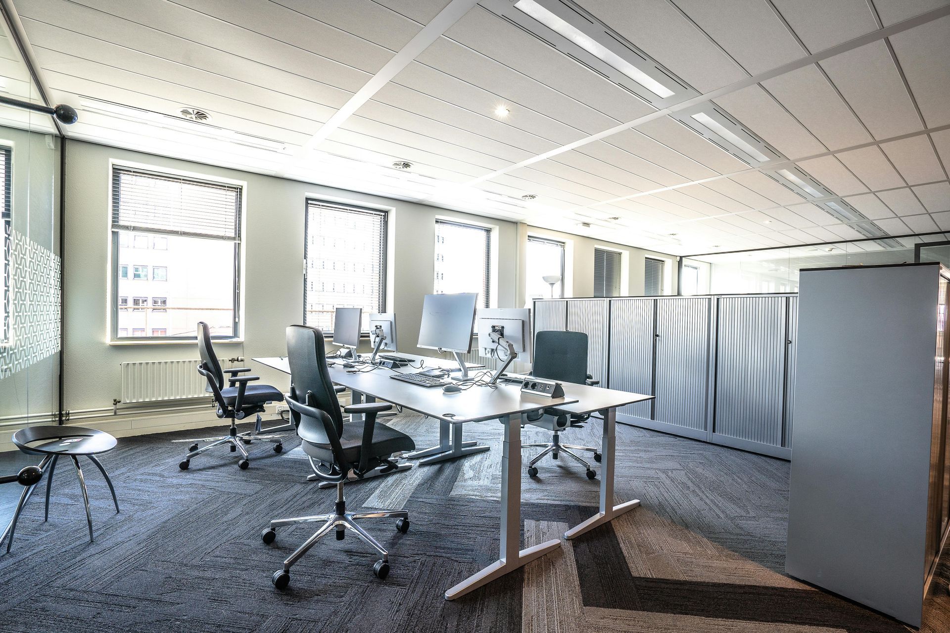 Empty modern office with desks, rolling chairs, computers, and partition screens under bright windows