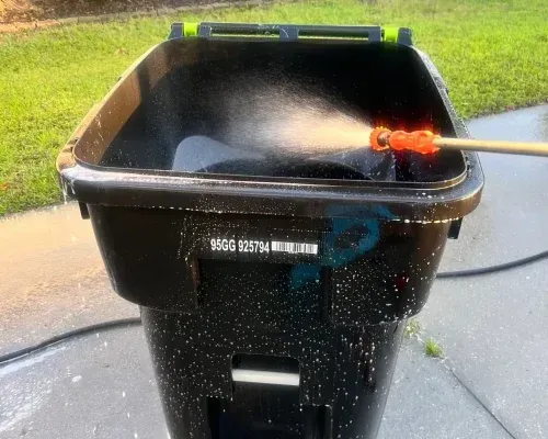 Cleaning a black trash can with a pressure washer outside.