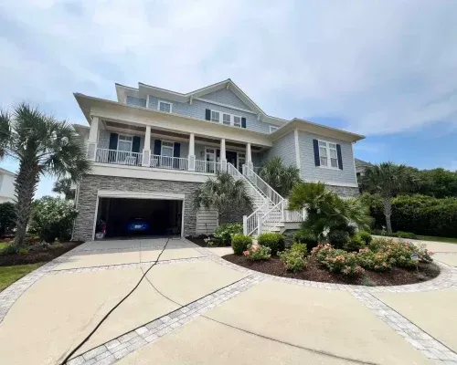 Two-story blue house with garage and porch, surrounded by palm trees and landscaping, on a sunny day.