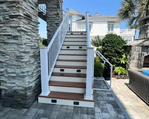Staircase with white railing, brown steps, and integrated lights, leading up to a house on a sunny day.
