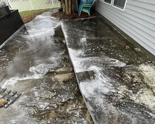 Concrete patio being cleaned with soap and water, near a house and a tree, creating foamy white suds.