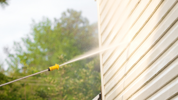 A person power washes a house siding with a yellow nozzle, removing dirt outdoors.