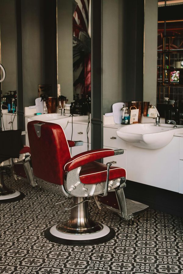 Red barber chair in a barbershop with mirrors, sink, and various products. Patterned floor.