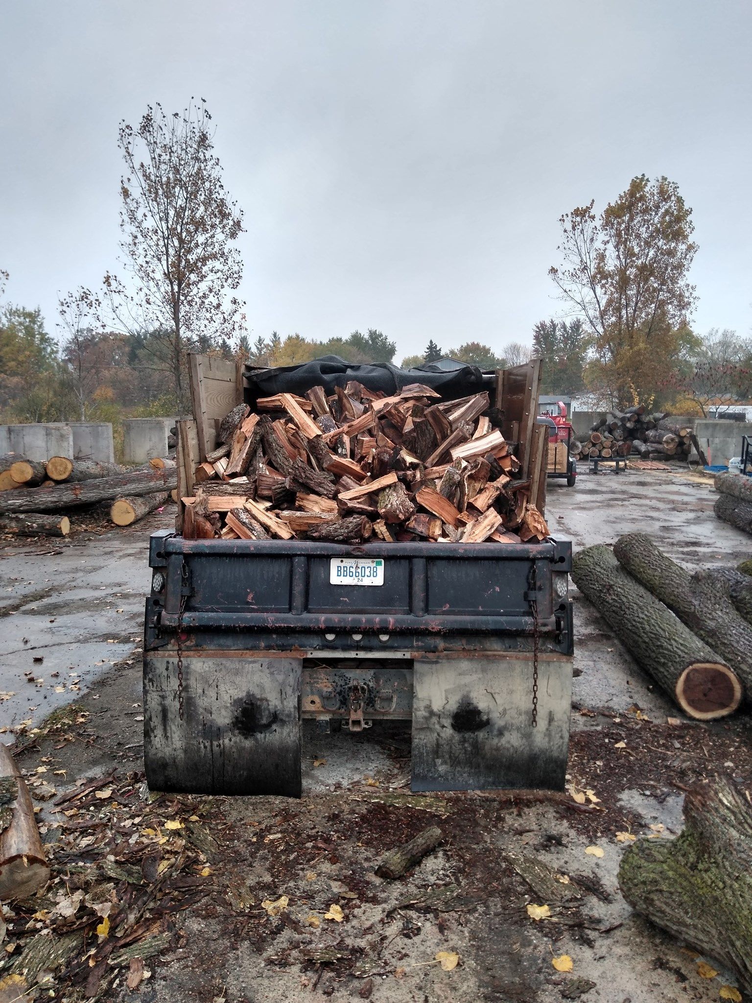 Truck bed filled with split firewood, parked in a lumber yard on a cloudy day.