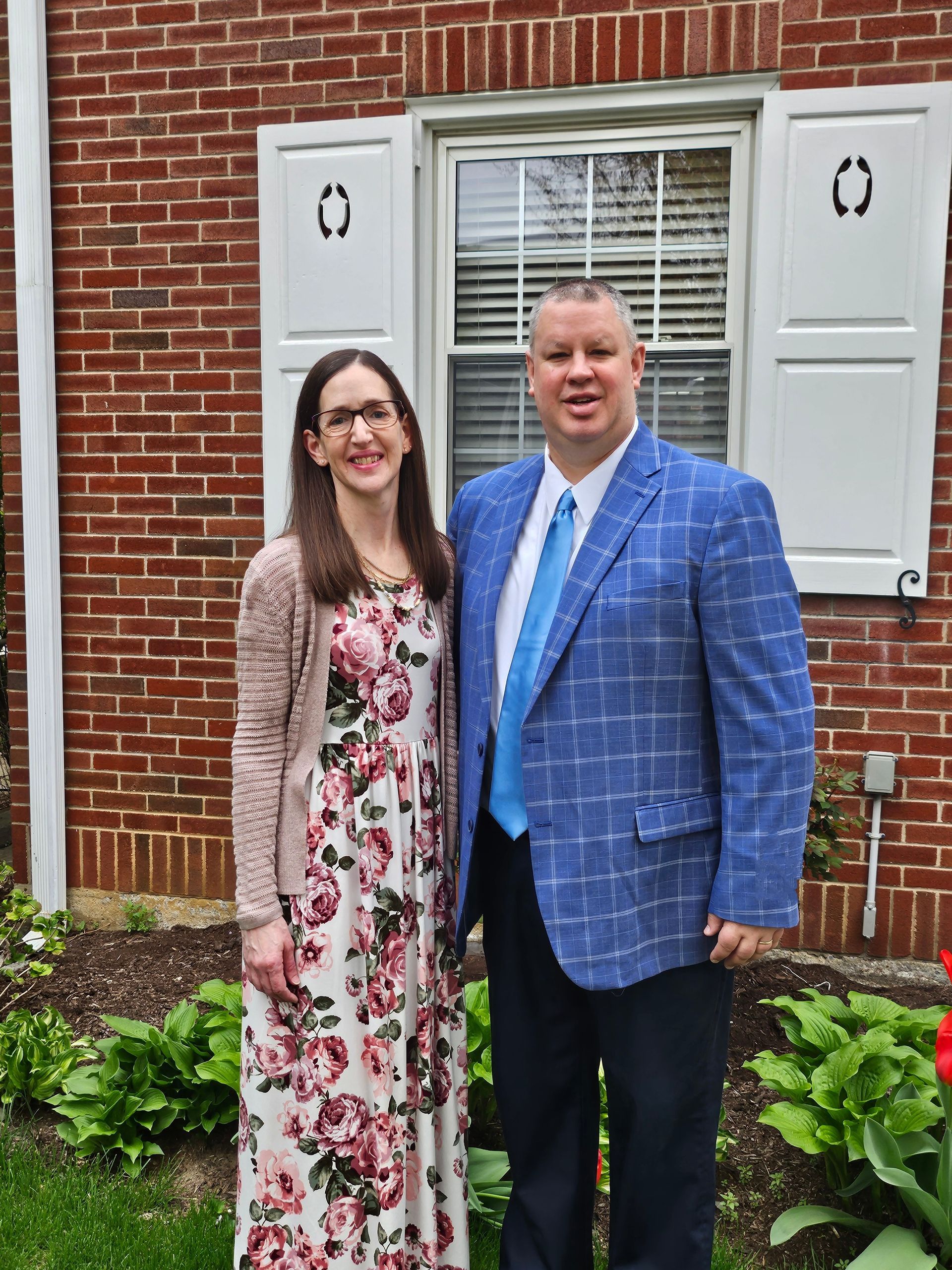 Woman and man stand in front of flowering tree, woman in blue dress and jacket, man in suit.