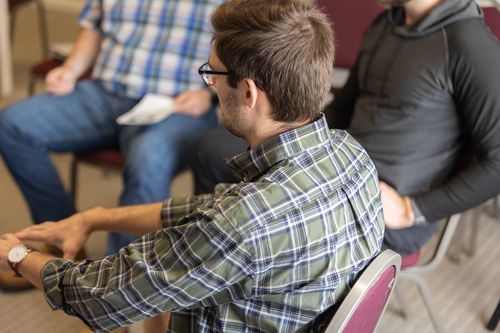 Man in plaid shirt, glasses, and watch, sitting in a circle with two other men.