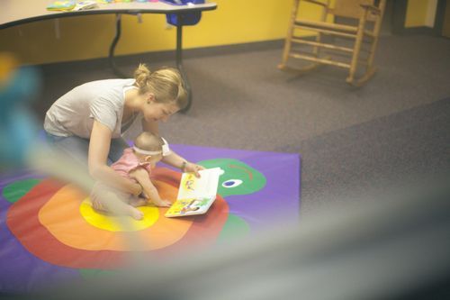 Woman reads to a baby on a colorful mat in a room with a rocking chair.