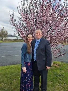 Woman and man stand in front of flowering tree, woman in blue dress and jacket, man in suit.