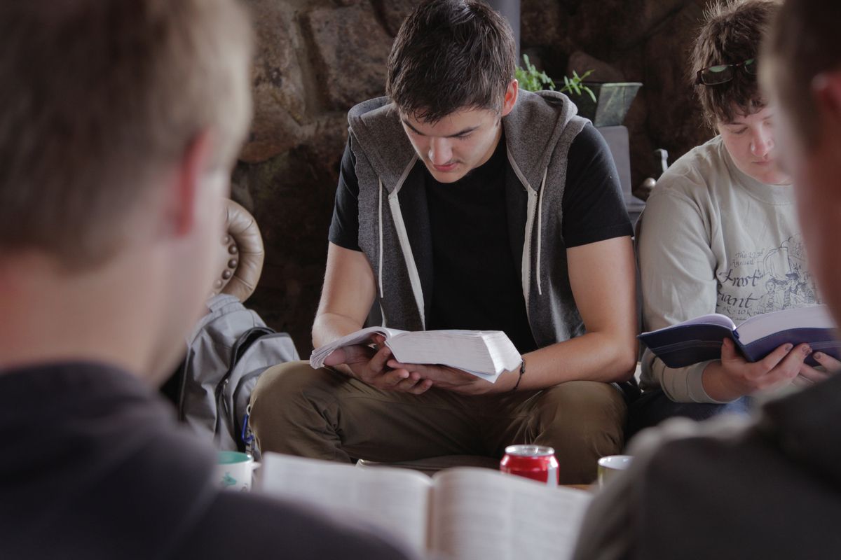 Group of young men studying books, one holding a white container, in a rustic indoor setting.