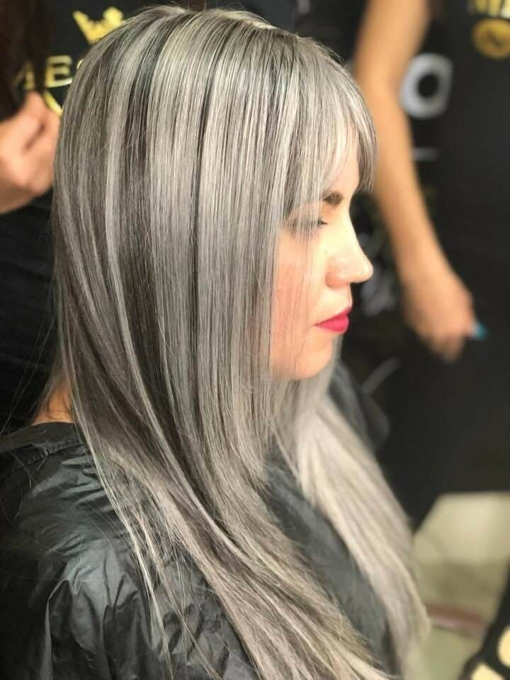 Mujer con cabello gris, liso y flequillo, con capa en un salón de belleza, mirando hacia un lado.