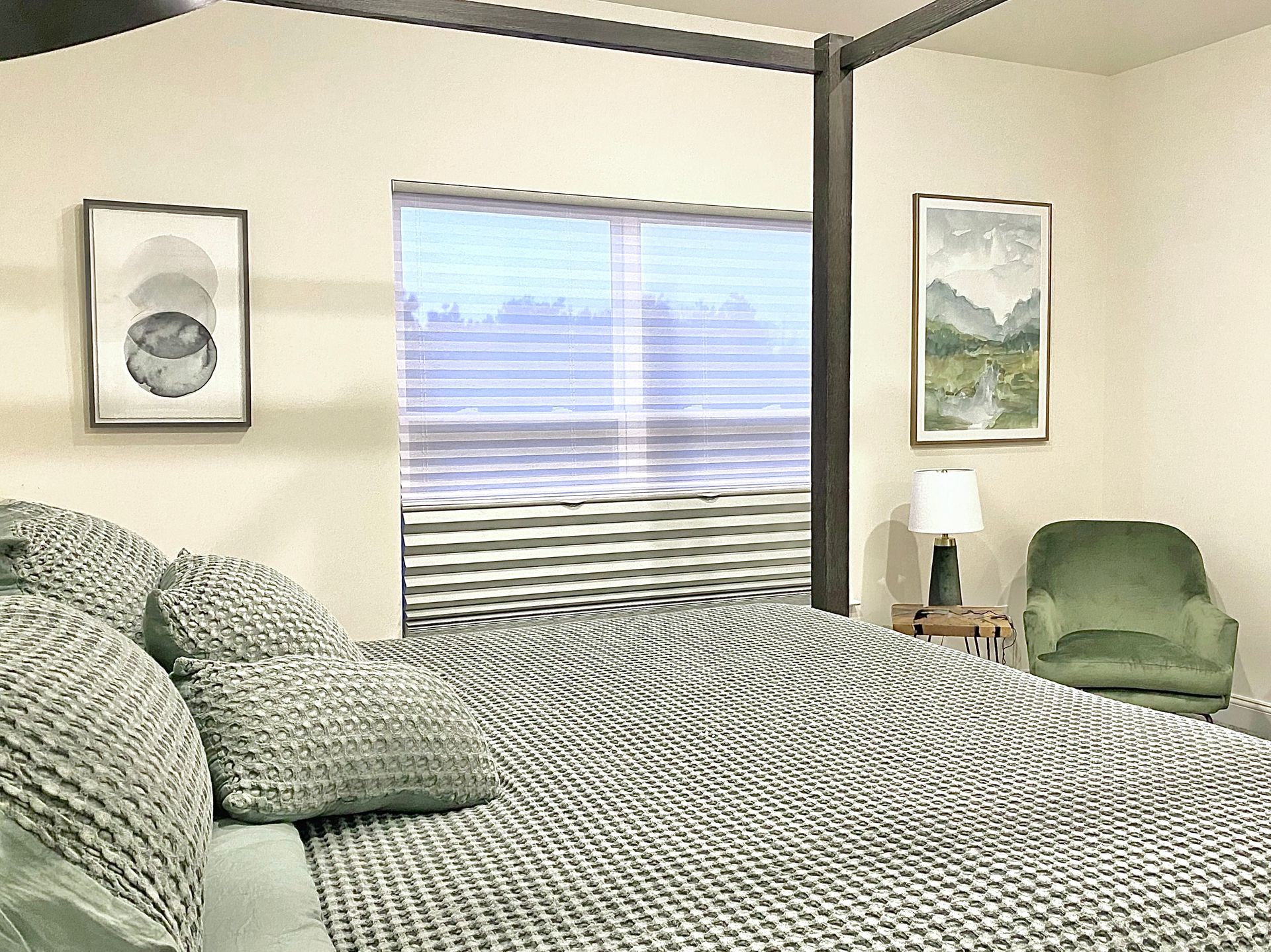 Bedroom with a bed covered in a patterned blanket, artwork, window with blinds, and a green armchair. By Robin Bradley Designs 