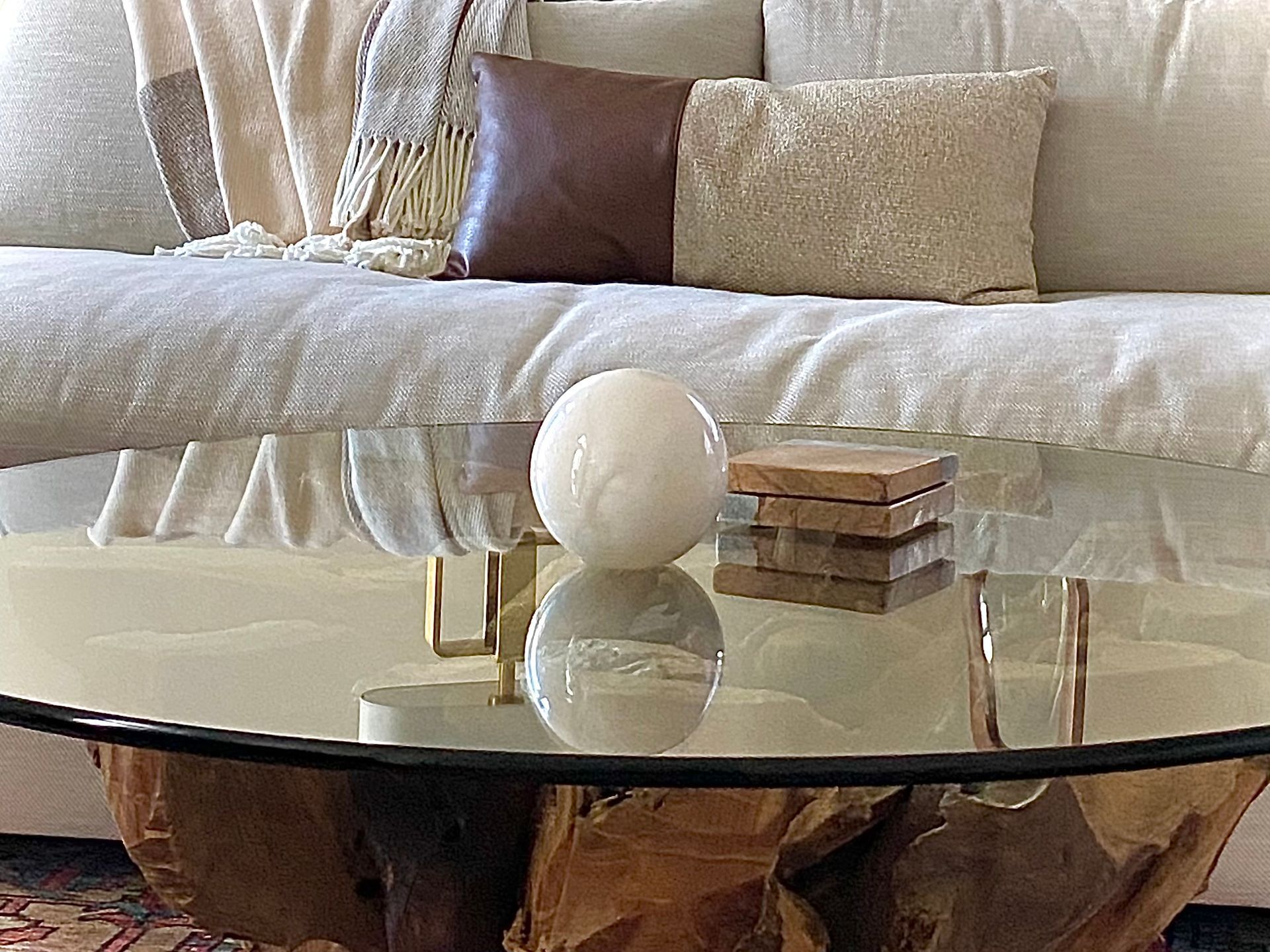 A glass coffee table with a white orb and coasters in front of a beige sofa with pillows. By Robin Bradley Designs