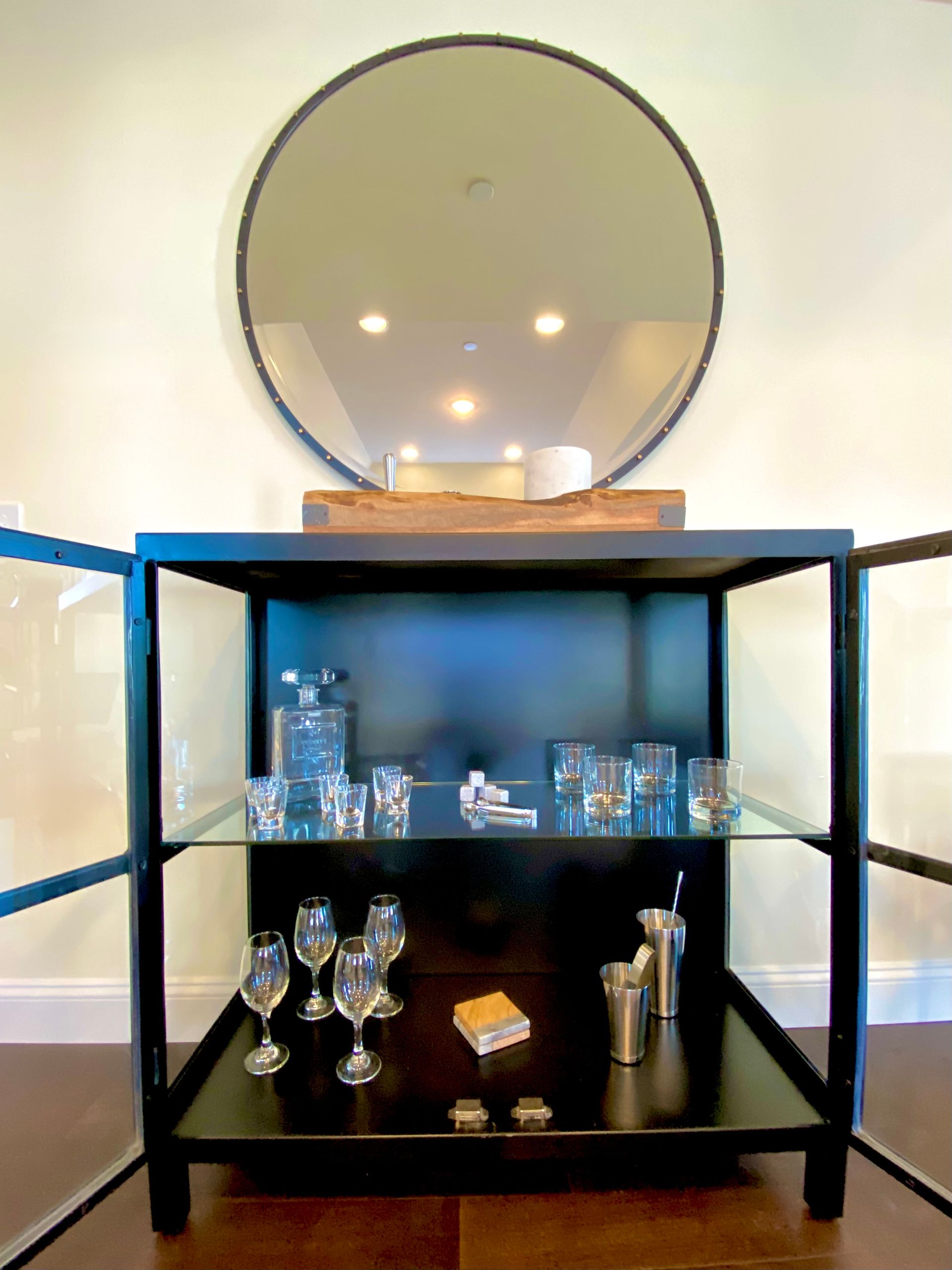 Black bar cabinet with glassware, a large round mirror, and wooden shelf on a hardwood floor. By Robin Bradley Designs 