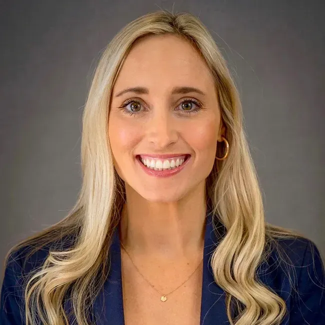 Blonde woman with a big smile, wearing a navy blazer and gold jewelry, against a gray background.