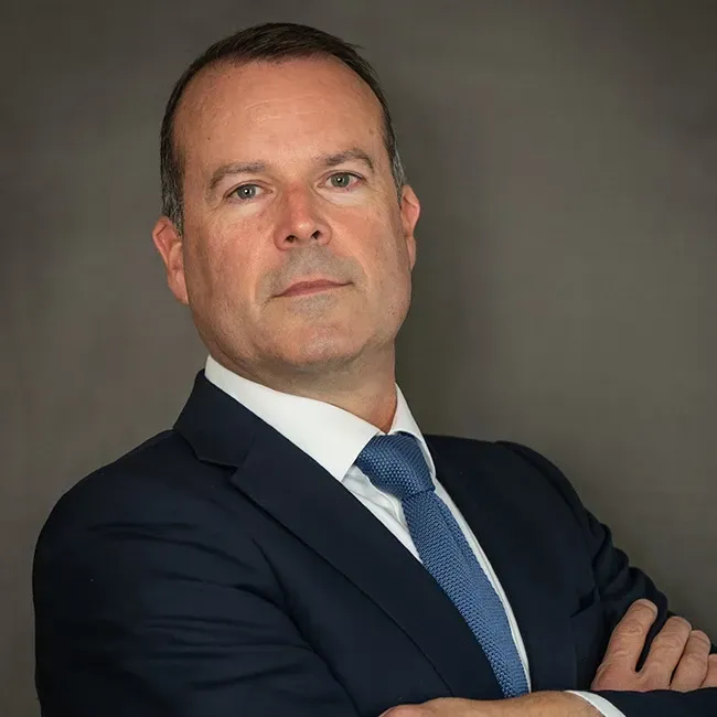 Man in a suit with arms crossed, looking forward with a serious expression, against a gray backdrop.