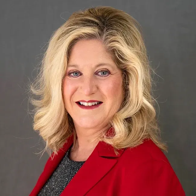 Woman with blonde hair, wearing a red blazer over a black and gray top, smiling.