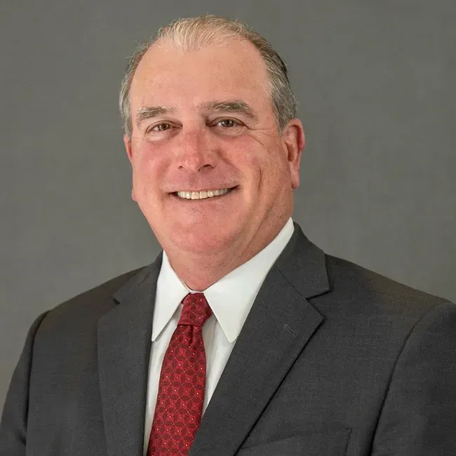 Man in suit and red tie smiling, headshot against a gray background.