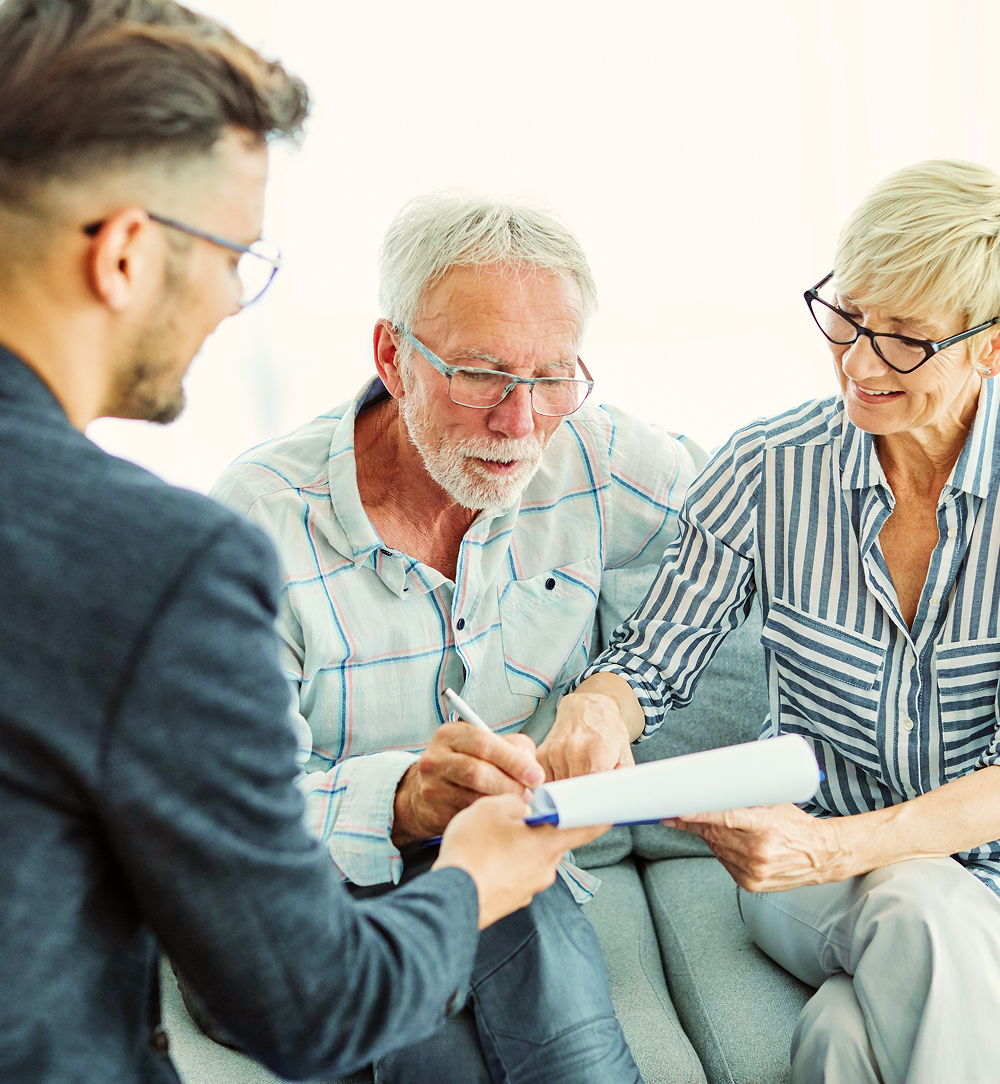 Man in suit hands document to older man, who signs it, while older woman looks on.